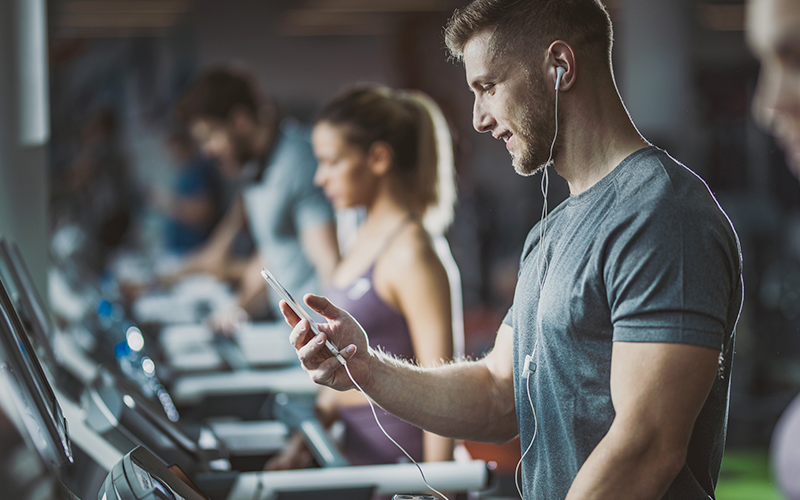 Mann steht mit Handy und Kopfhörer auf dem Laufband